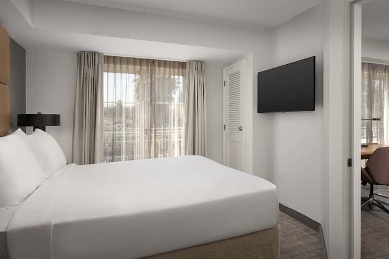 Contemporary hotel room featuring queen bed, wall-mounted TV, glass doors to balcony, and neutral color palette