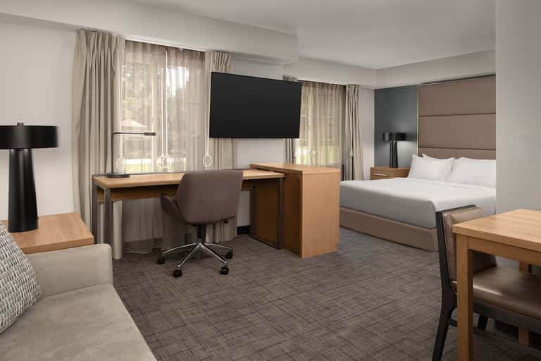 Spacious hotel suite with bed, work desk, chair, and flat-screen TV overlooking windows with palm trees visible outside