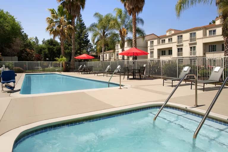 Resort pool area with two pools, red umbrellas, lounge seating, palm trees, and residential buildings overlooking the courtyard
