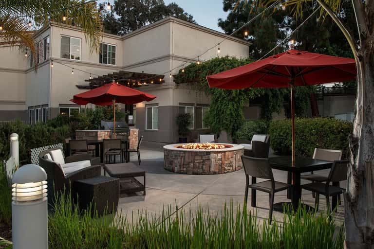 Outdoor patio with fire pit, lounge seating, red umbrellas, dining area, and landscaped gardens at dusk with ambient lighting