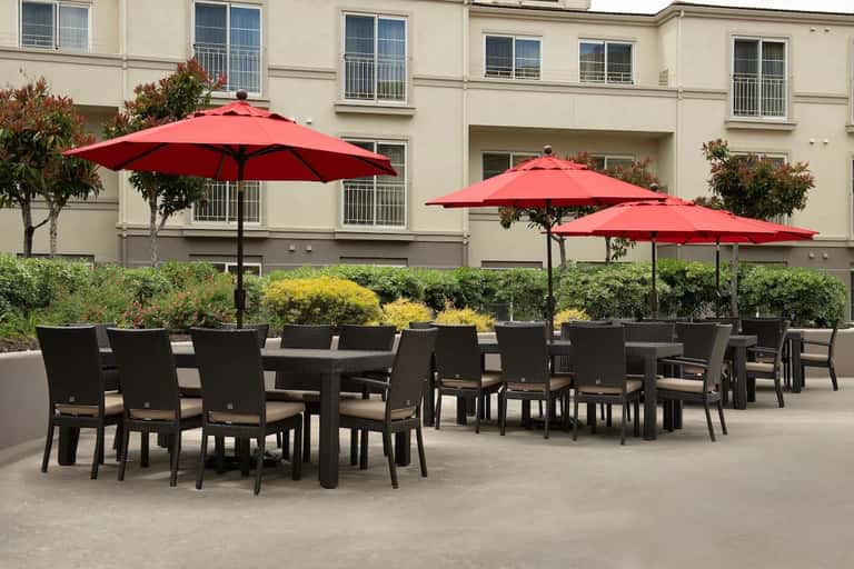 Outdoor courtyard dining area with black wicker chairs, tables, red umbrellas, and multi-story residential building facade