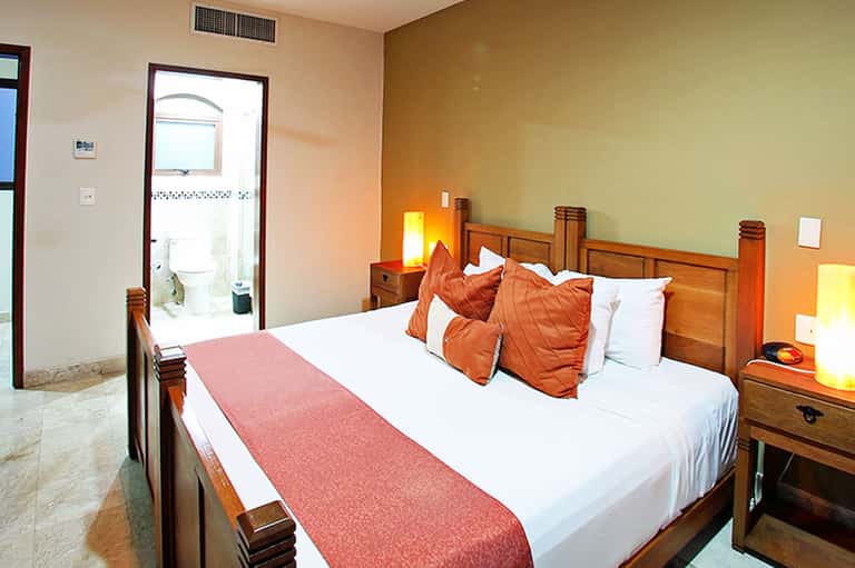 Hotel bedroom with wooden bed frame, orange pillows, white bedding, and ensuite bathroom