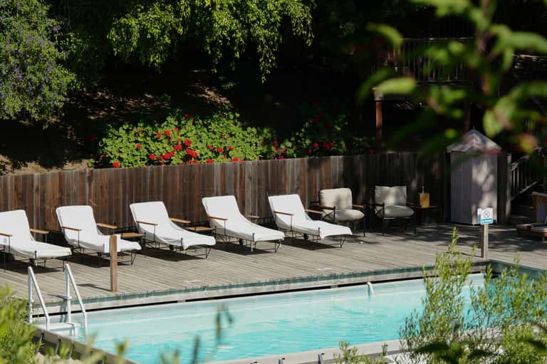 Outdoor pool deck with white lounge chairs, wooden fence, and red flowers under trees