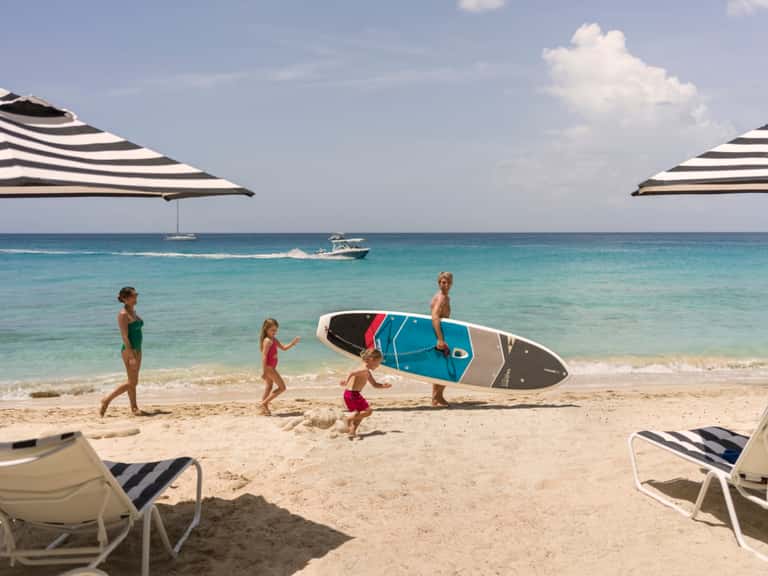 Family on sandy beach with striped umbrellas, paddleboards, and clear turquoise ocean water