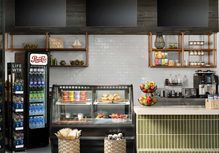 Modern cafe counter with white subway tile, wooden shelving, Pepsi cooler, pastry display case, and fresh fruit bowls