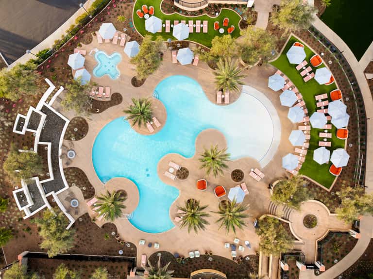 Aerial view of resort pool complex with curved pools, pink lounge chairs, umbrellas, and surrounding palm trees