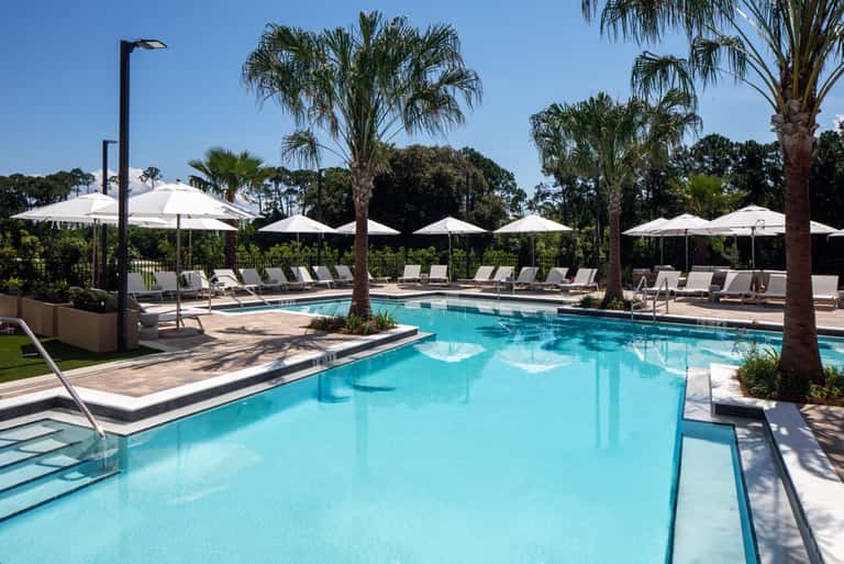 Large resort pool surrounded by palm trees, white umbrellas, and lounge seating areas