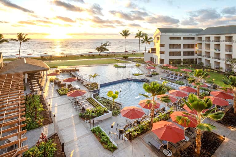 Beachfront resort at sunset with pool, red umbrellas, palm trees, and ocean view