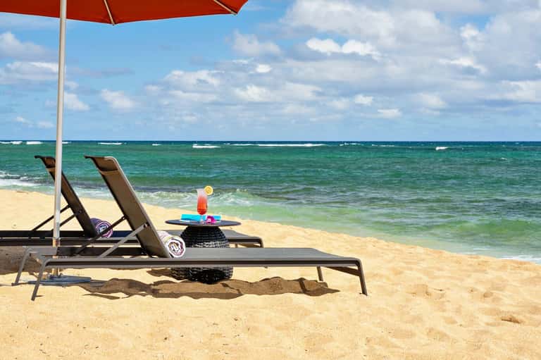 Beach lounge chairs under red umbrella with tropical cocktail, overlooking turquoise ocean waters
