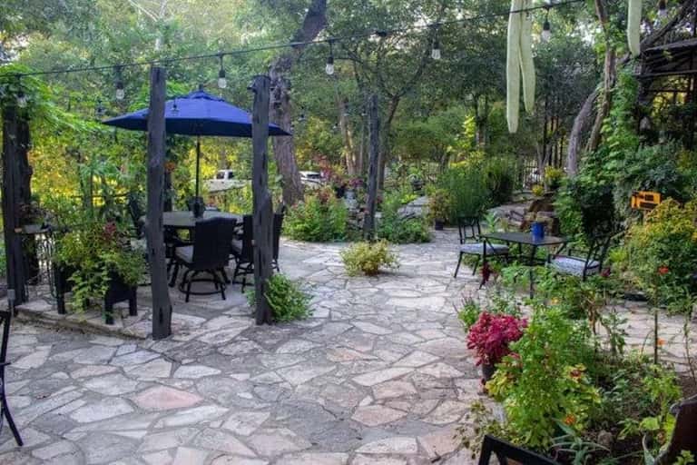Stone patio pathway through lush forest with pergola, dining area, and garden plantings