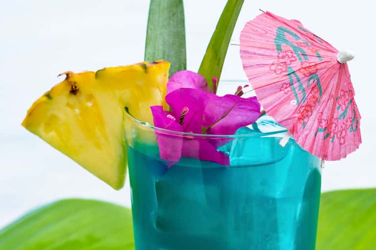 Tropical cocktail with pineapple slice, pink umbrella, magenta flowers in turquoise glass with green straws