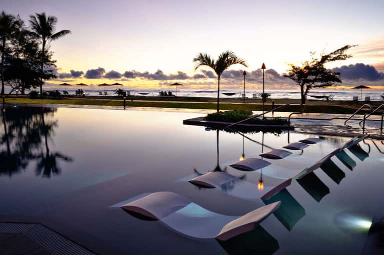 Illuminated lounge chairs reflected in still water at dusk with palm trees and sunset