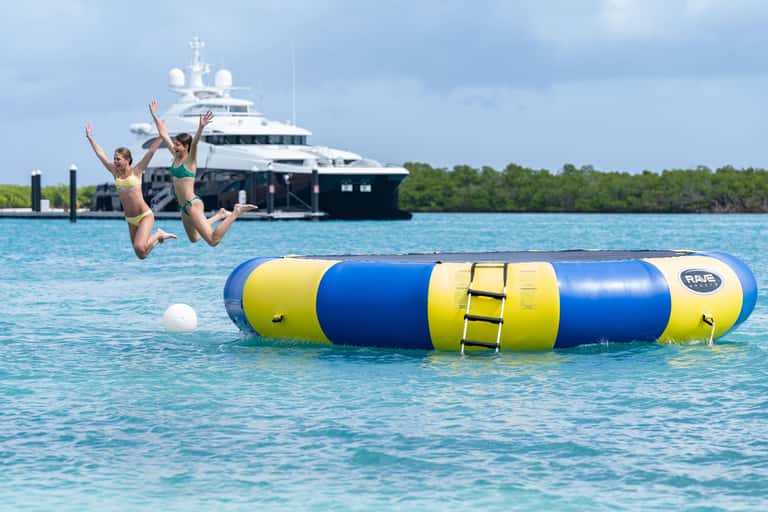 Two children jumping off inflatable water toy with yacht and mangroves in background