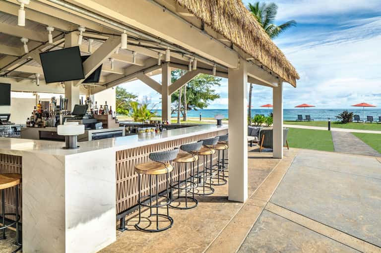 Beachfront bar with thatched roof, white marble counter, wooden stools, ocean view and red umbrellas