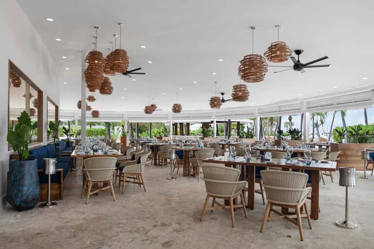 Beachfront restaurant with wooden tables, woven pendant lights, and panoramic ocean views through windows