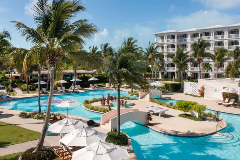 Tropical resort pool area with loungers, umbrellas, palm trees, and white hotel building