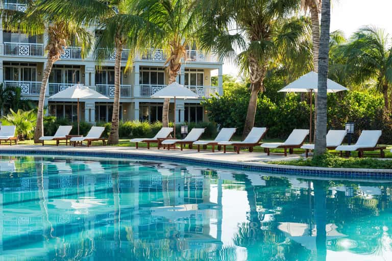 Resort pool with white loungers, umbrellas, and palm trees beside colonial-style white building