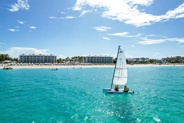 Sailboat with two people on turquoise ocean near sandy beach with resort buildings