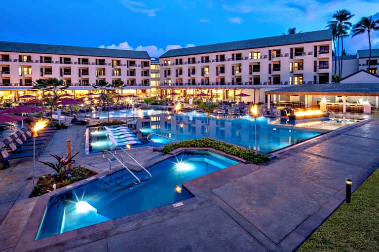 Illuminated modern resort pool at dusk with pink building, fire pits, and lounge chairs