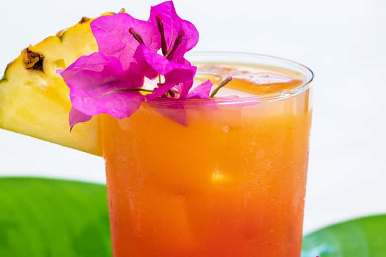 Tropical cocktail with orange color garnished with pineapple and magenta bougainvillea flower