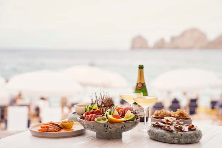 Beachside dining setup with seafood platters, champagne, and coastal view with white umbrellas