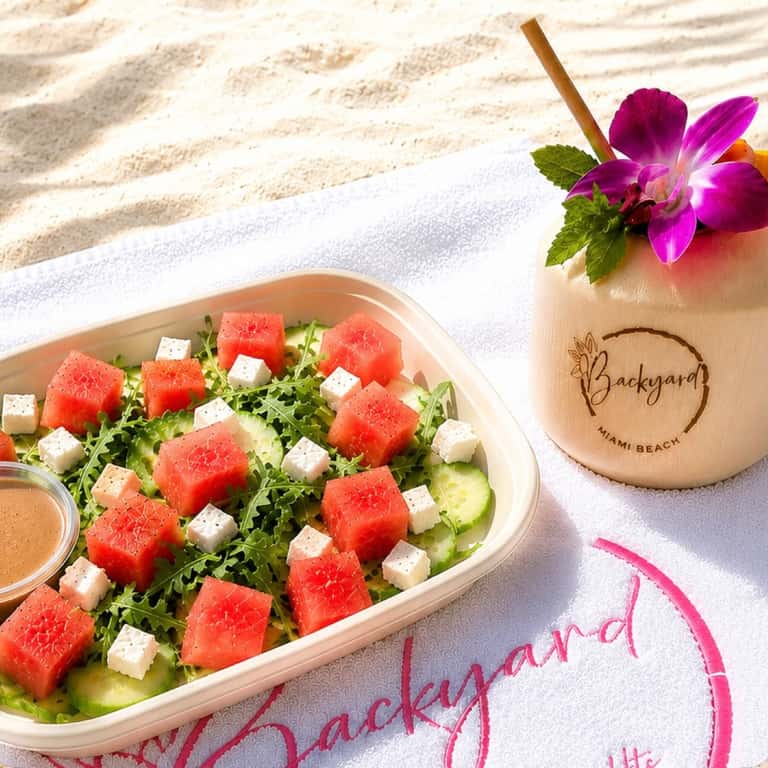 Watermelon and feta salad with tropical drink on beach towel at Backyard Miami Beach