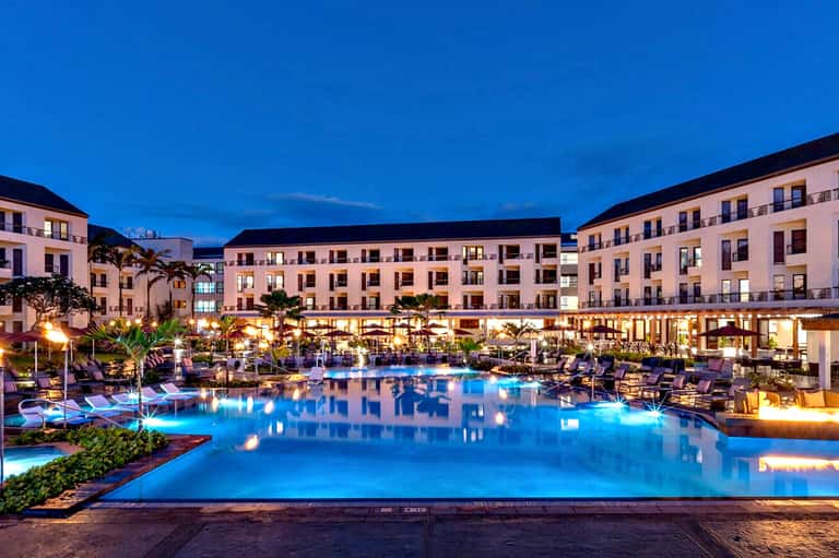 Illuminated resort pool at dusk with multi-story hotel buildings and palm trees
