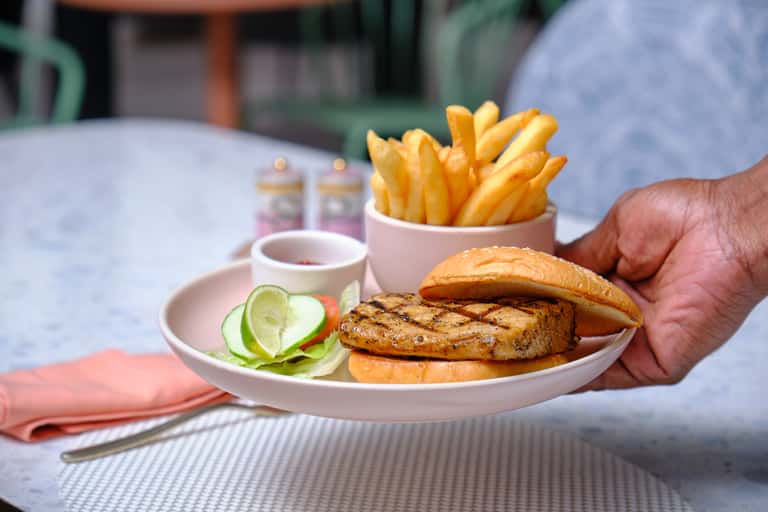 Server holding grilled chicken burger with fries and cucumber slices at outdoor dining table