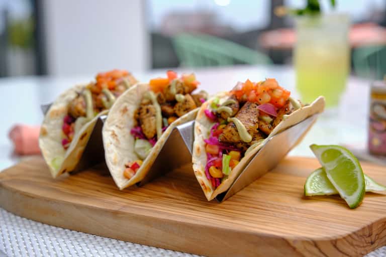 Three fish tacos with colorful toppings on wooden board with lime wedges and beverage