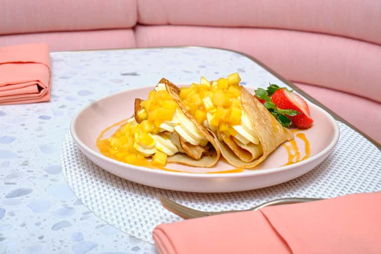 Crepes with mango compote, banana, and strawberry on pink plate with orange drizzle