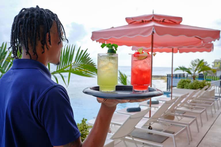 Server in blue uniform holding tray with tropical cocktails on beachfront patio with red umbrellas and ocean view