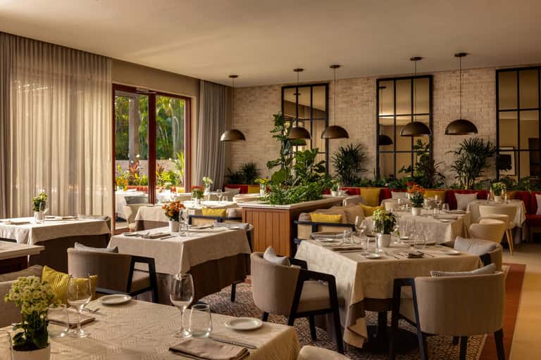 Fine dining restaurant with beige upholstered chairs, potted plants, brick walls, and garden views