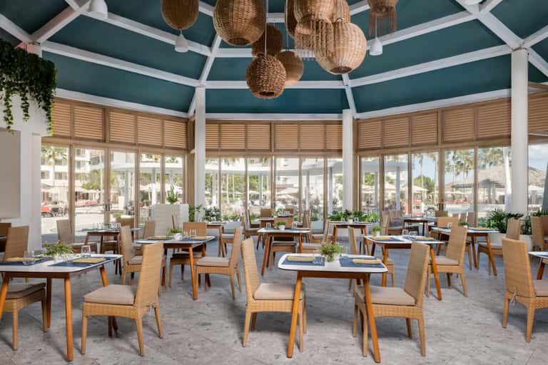 Modern restaurant dining room with teal ceiling, woven pendant lights, wooden chairs, and floor-to-ceiling windows overlooking a beachfront promenade.
