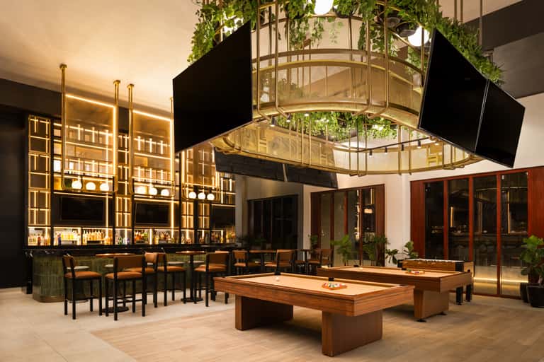 Luxury lounge with gold-accented bar, billiard tables, hanging plants, and ambient lighting in a contemporary indoor setting.