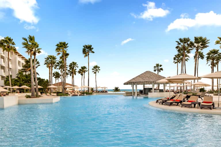 Resort pool with crystal-blue water, palm trees, thatched umbrellas, lounge chairs, and ocean views under clear blue sky.