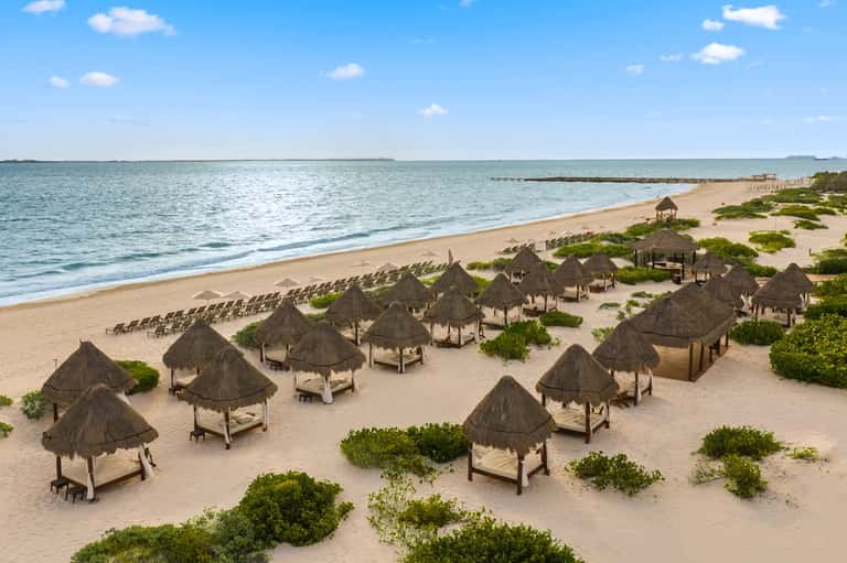 Aerial view of pristine beach with rows of thatched-roof cabanas, white sand, calm turquoise waters, and coastal vegetation.