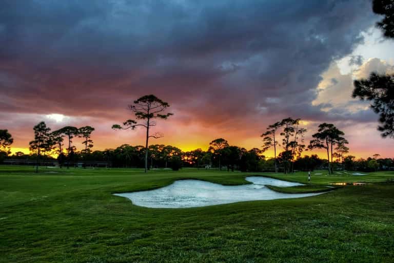 Golf course at sunset with sand bunkers, tall pine trees, and dramatic colorful sky