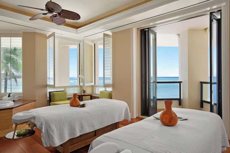 Beachfront spa treatment room with ocean views, ceiling fan, and two massage beds
