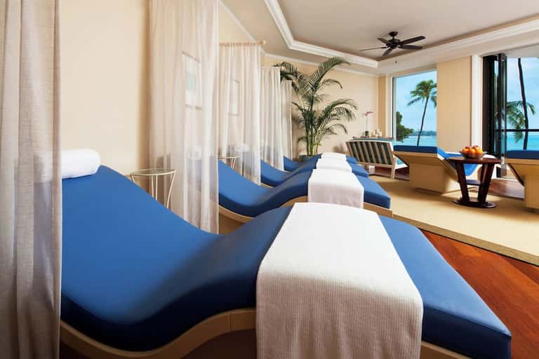 Spa treatment room with multiple massage beds, ocean view, and tropical plant décor