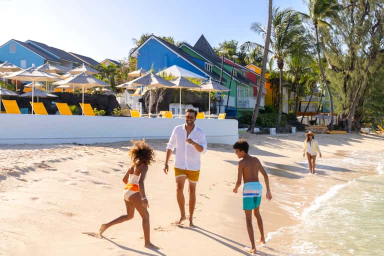 Family playing on sandy beach with colorful beachside buildings and palm trees
