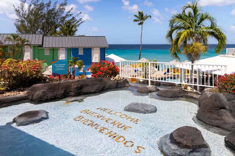 Beachfront welcome sign with colored cottages, palm trees, and turquoise ocean water in background