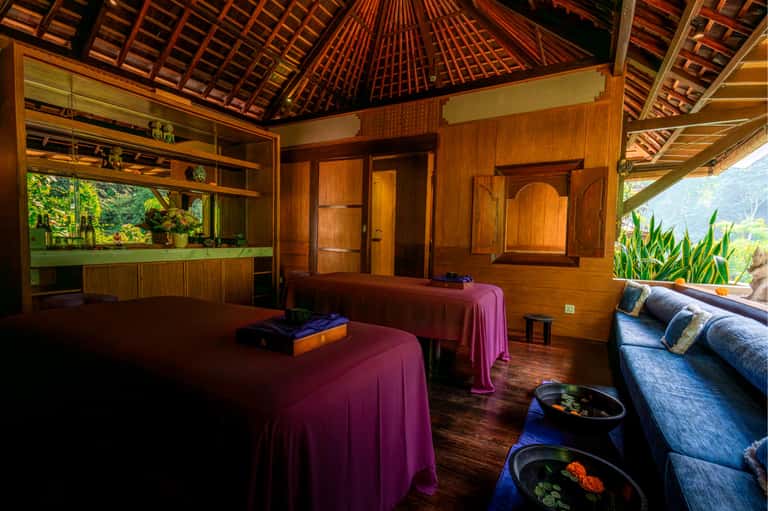 Wooden spa treatment room with high ceiling, massage tables with pink linens, and jungle views