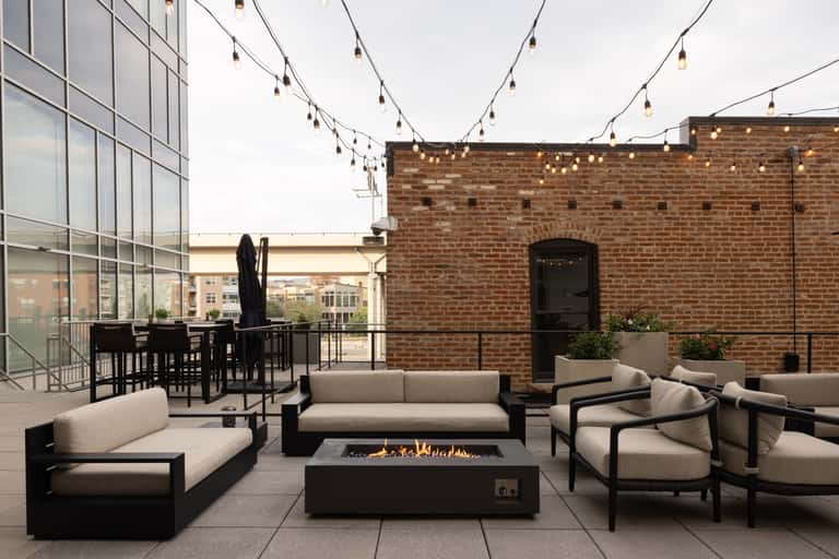 Rooftop lounge with beige seating, fireplace, string lights, and brick wall overlooking city