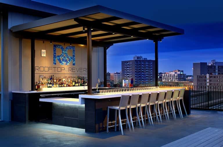 Rooftop bar at dusk with illuminated pergola, full-service bar, and city skyline backdrop