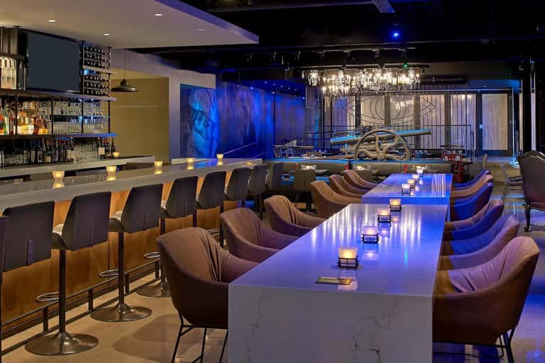 Modern bar and lounge with blue mood lighting, marble countertop, and floor-to-ceiling windows at night