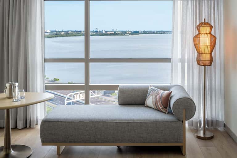 Modern hotel suite with gray daybed, large windows, and panoramic water and cityscape views