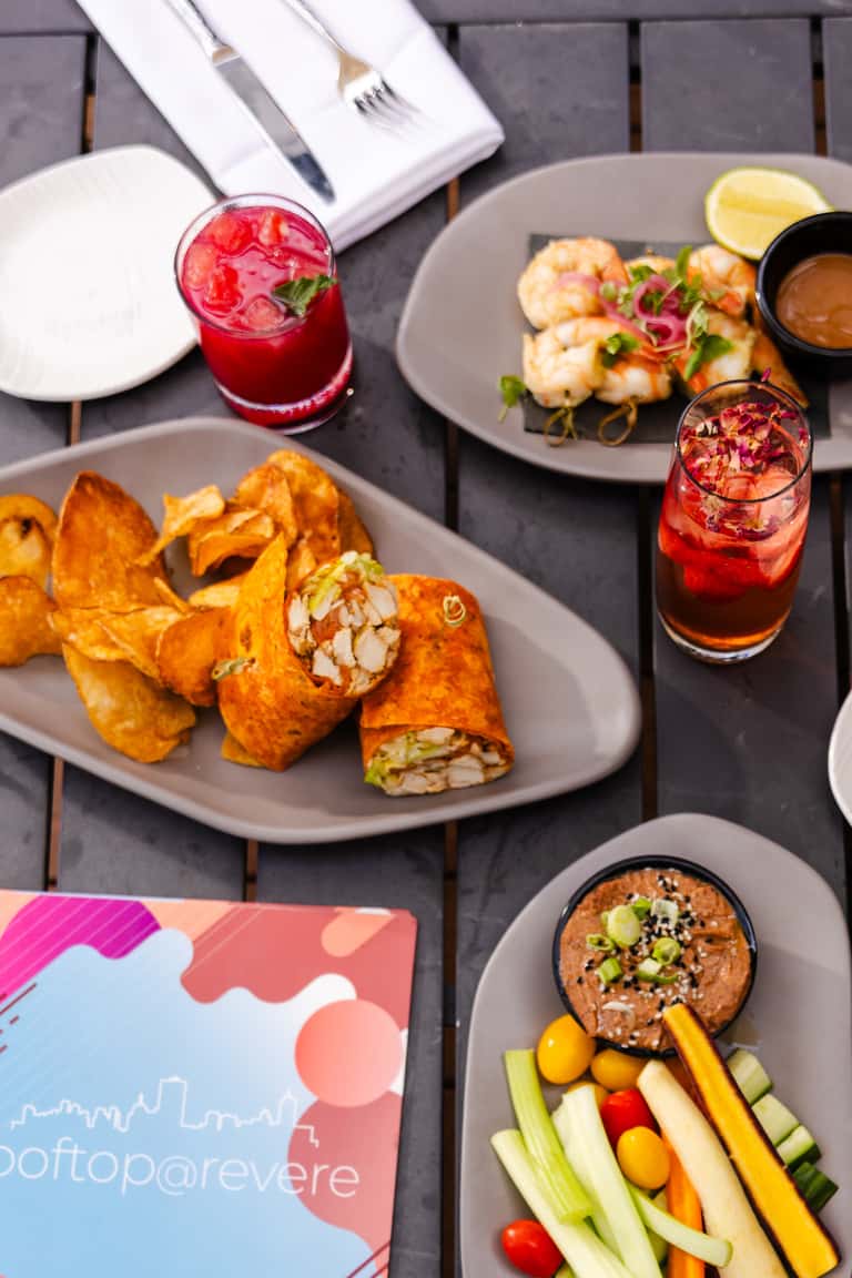 Overhead view of gourmet appetizers, colorful cocktails, and white place settings on dark table