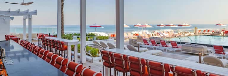 Resort poolside bar with red lounge chairs overlooking ocean and beach umbrellas