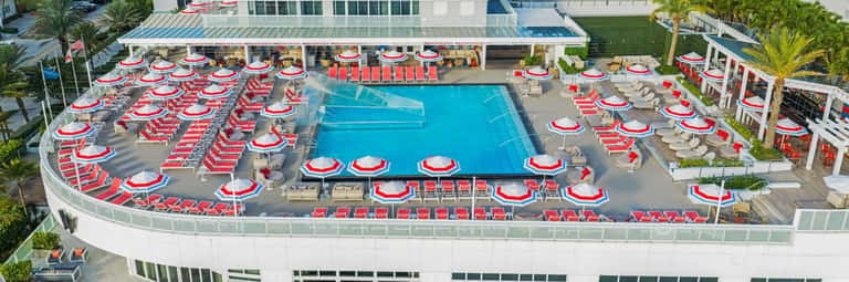 Aerial view of resort pool deck surrounded by red and white umbrellas, lounge chairs, and palm trees