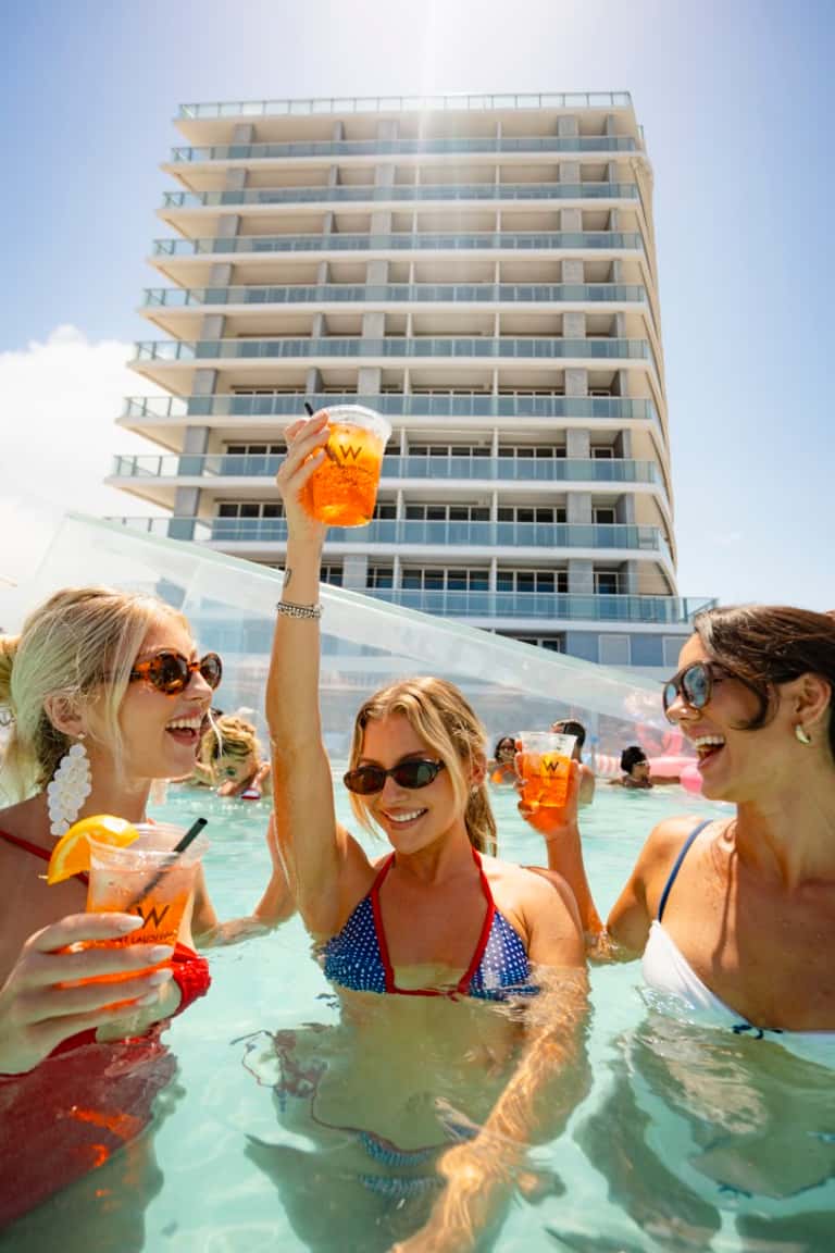 Women in a pool holding colorful drinks in front of a tall beachfront hotel building
